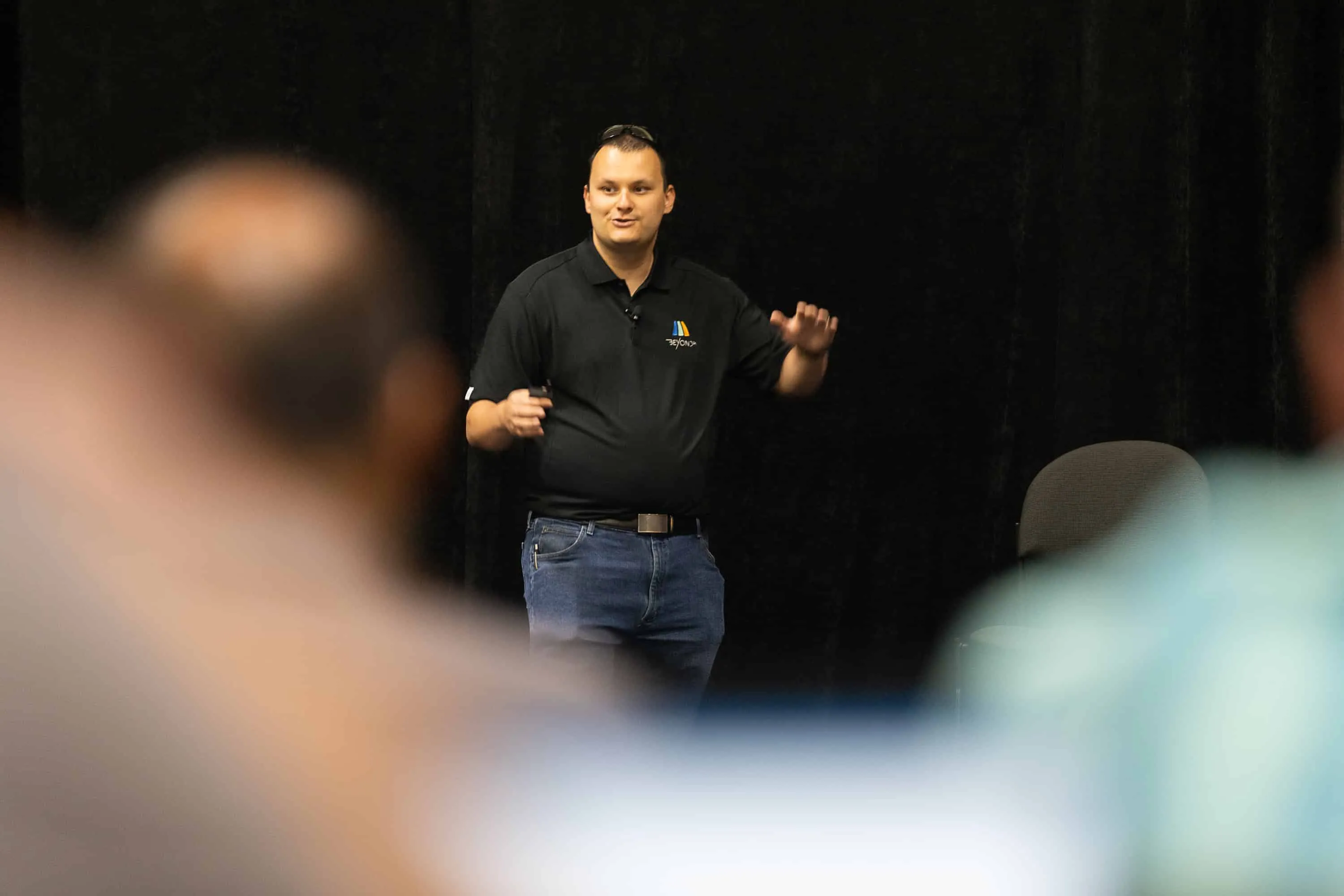Dom Kirby speaking on stage in front of a seated audience, gesturing mid-talk with a clicker in hand